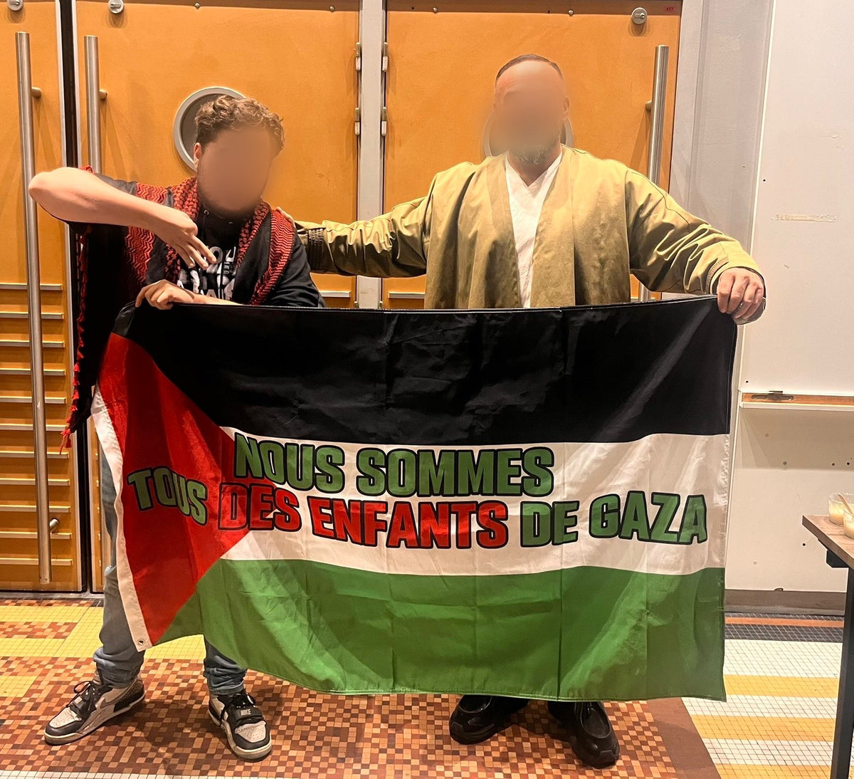 Deux personnes présentent un drapeau personnalisé portant l’inscription « Nous sommes tous des enfants de Gaza ».